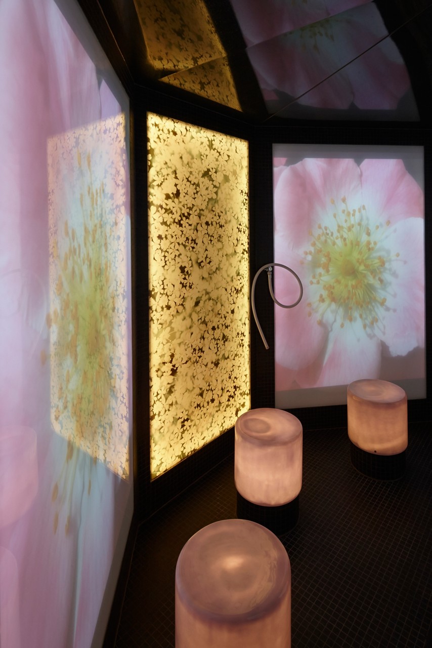 Three glowing cylindrical stools sit amid illuminated floral panels and projected flower images; a curved shower wand hangs on the wall in a dim, tiled spa-like room.